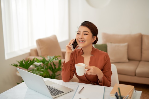 Mujer de negocios asiática joven que sostiene la taza y que usa el teléfono inteligente mientras está sentado en el escritorio de la oficina con la computadora portátil