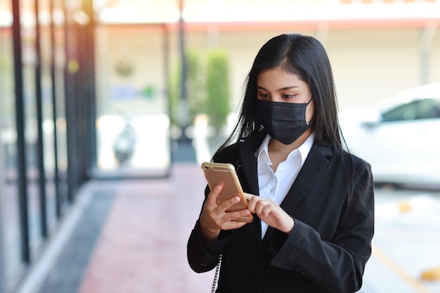 Mujer de negocios asiática joven en traje negro de negocios con máscara de protección para el cuidado de la salud caminando en público al aire libre y con smartphone. Nuevo concepto de distanciamiento normal y social