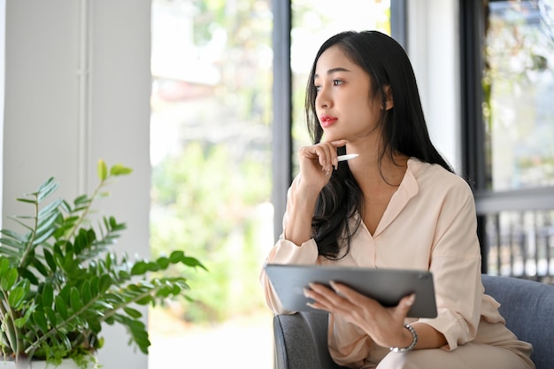 Una mujer de negocios asiática milenaria pensativa reflexionando sobre mirar por la ventana usando una tableta