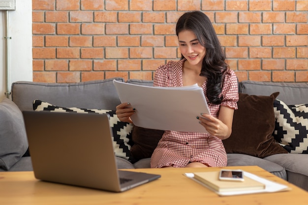 Mujer de negocios asiática mirando sus planes de trabajo frente a su computadora portátil en su sala de estar