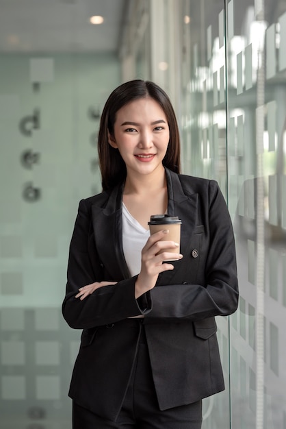 Mujer de negocios asiática de pie sosteniendo un café en una oficina moderna.