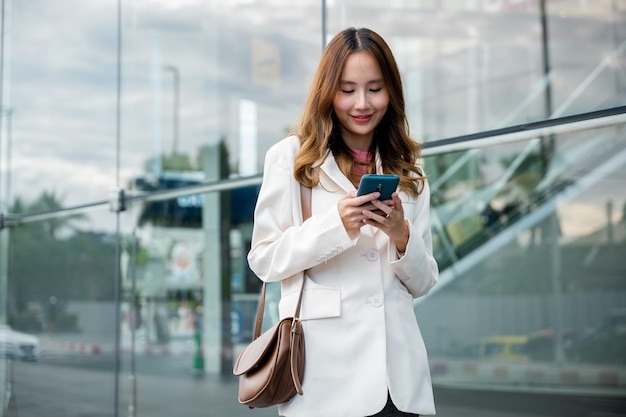 Mujer de negocios asiática que trabaja con un teléfono móvil de pie contra el edificio de la calle cerca de la oficina, Retrato mujer de negocios exitosa sonriendo sosteniendo el chat de aplicaciones de uso de teléfonos inteligentes en línea por la mañana