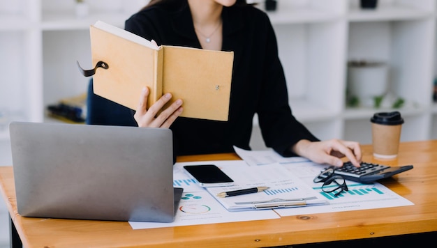 Mujer de negocios asiática que usa una computadora portátil y trabaja en la oficina con un documento de calculadora en el escritorio haciendo planificación analizando el concepto de análisis de finanzas de inversión del plan de negocios del informe financiero