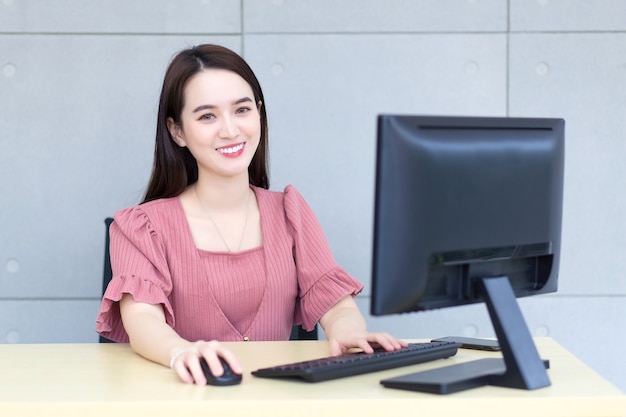 Mujer de negocios asiática que usa un vestido rosa está trabajando mientras se sienta y sonríe en una oficina escribiendo