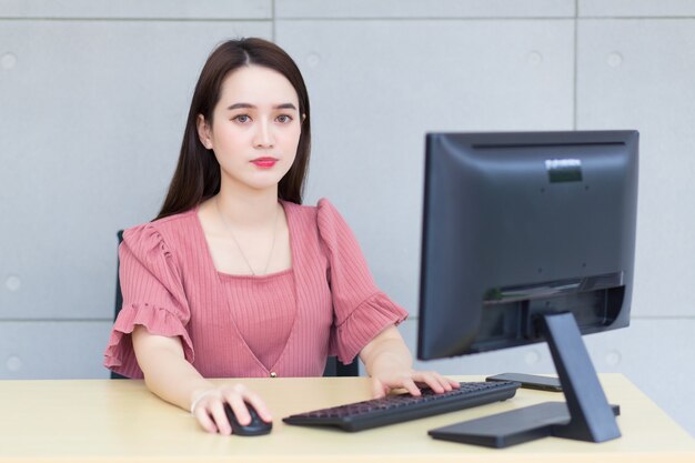 Mujer de negocios asiática que usa un vestido rosa trabajando en una oficina escribiendo en un teclado y un mouse.