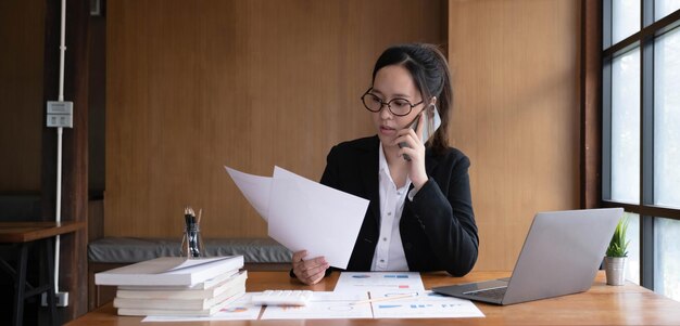Una mujer de negocios asiática seria que usa una computadora portátil mirando la computadora hablando por teléfono consulta al cliente sentada en el escritorio enfocada en la gerente de ventas que hace una llamada de negocios móvil para discutir el trabajo con el cliente en la oficina