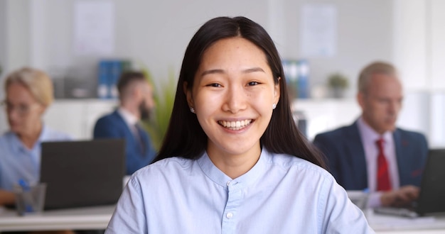 Una mujer de negocios asiática sonriente habla con una cámara web para hacer una videoconferencia sentada en una oficina de espacio abierto