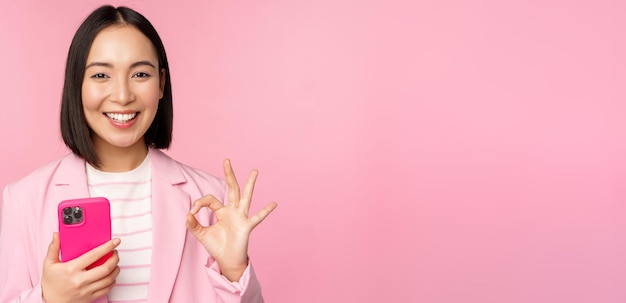 Mujer de negocios asiática sonriente que muestra un signo de aprobación mientras usa una aplicación de teléfono móvil recomendando una aplicación de teléfono inteligente de pie sobre un fondo rosa