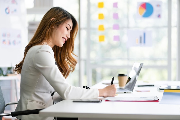 Mujer de negocios asiática sonriente sentada y escribiendo ideas de negocios en la nota de papel.