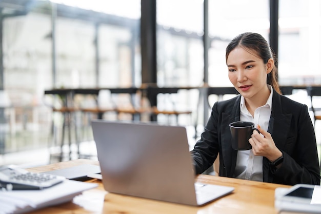 Mujer de negocios asiática con traje formal en la oficina sosteniendo una taza trabajando en una laptop en la oficina