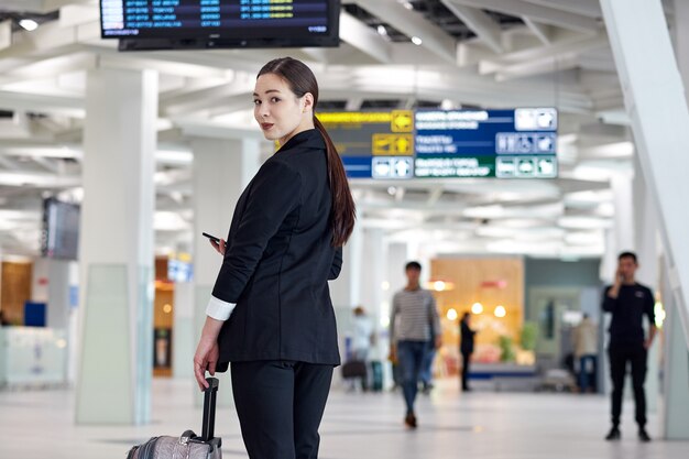 Mujer de negocios asiáticos en el aeropuerto con bolsa de tranvía, cerca de la pantalla de vuelo buscando horario de vuelo