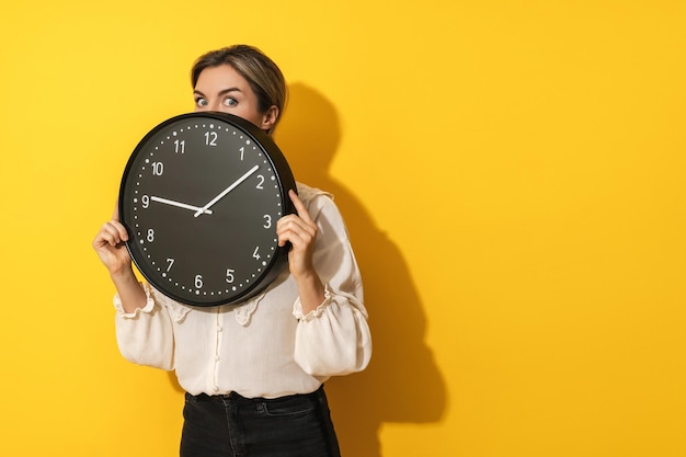 Mujer de negocios asomándose por detrás de un gran reloj en un fondo amarillo