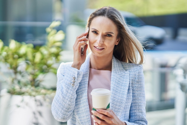 Mujer de negocios atractiva adulta con smartphone y café caminando en la ciudad. Foto de alta calidad