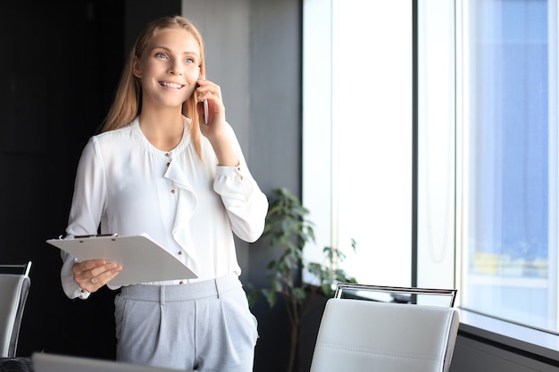 Mujer de negocios atractiva hablando con colegas por teléfono móvil mientras está de pie cerca de la ventana en la oficina.