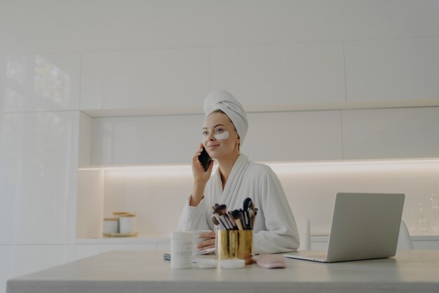 Mujer de negocios atractiva joven en albornoz y turbante de toalla en la cabeza con parches cosméticos debajo de los ojos hablando por teléfono móvil y trabajando en la computadora portátil mientras se prepara para trabajar en la mañana en casa