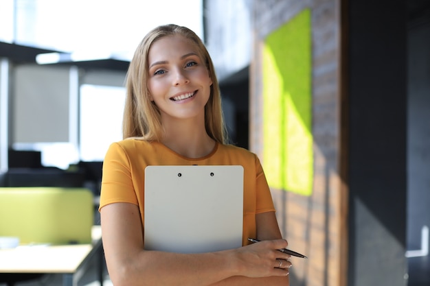 Mujer de negocios atractiva mirando a cámara y sonriendo mientras está de pie en la oficina.