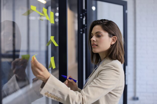 Foto mujer de negocios atractiva publicar pegatinas sobre vidrio en la oficina y escribir sobre ellos.