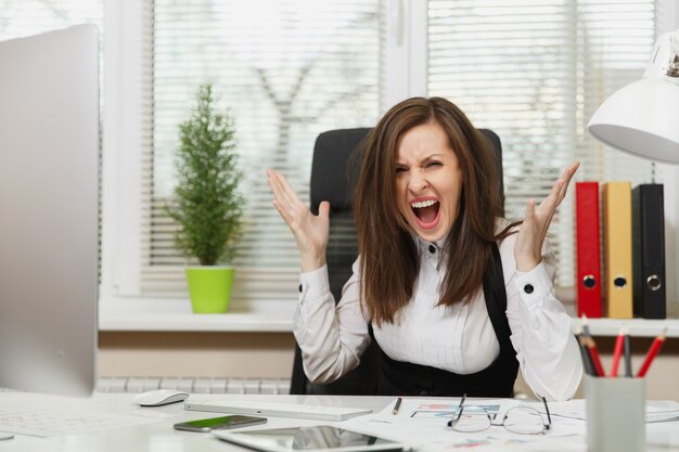 Mujer de negocios bastante enojada en traje sentada en el escritorio con tableta, trabajando en computadora con monitor moderno con documentos en oficina ligera, jurando y gritando, resolviendo problemas, concepto de emoción.