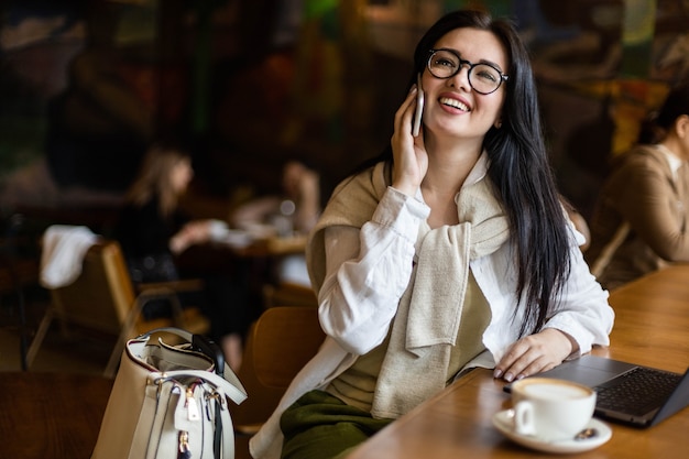 Mujer de negocios bastante joven hablando por teléfono inteligente sentado en la mesa de café con un portátil y una taza de café