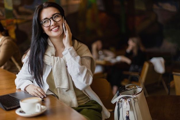 Mujer de negocios bastante joven hablando por teléfono inteligente sentado en la mesa de café con un portátil y una taza de café