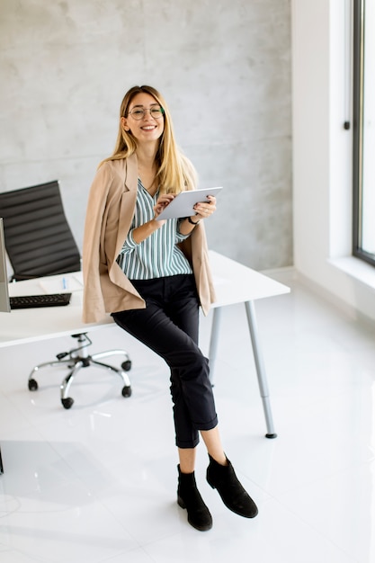 Foto mujer de negocios bastante joven que sostiene la tableta digital y de pie en la oficina moderna