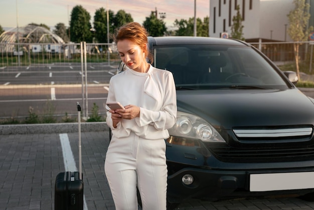 Mujer de negocios con bolsa de viaje usando teléfono móvil, reservando boletos de pie cerca del auto. concepto de viaje