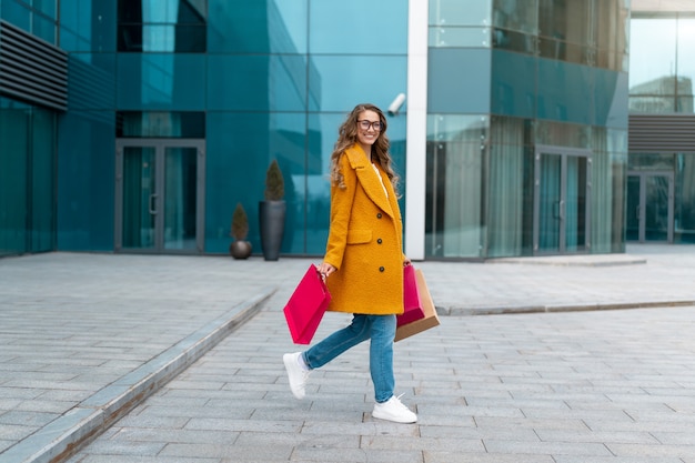 Mujer de negocios con bolsas de la compra vestida con abrigo amarillo caminando al aire libre Fondo de edificio corporativo Mujer caucásica Persona de negocios Edificio de oficinas Elegante empresaria Temporada Venta de longitud completa