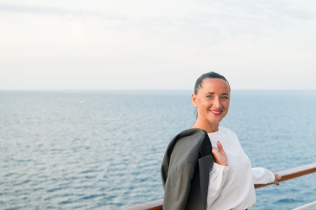 Mujer de negocios bonita o niña linda feliz con cara sonriente en blusa blanca, chaqueta gris, tiene manicura roja en la cubierta del barco viendo el mar, agua del océano sobre fondo de cielo nublado, viajes y turismo