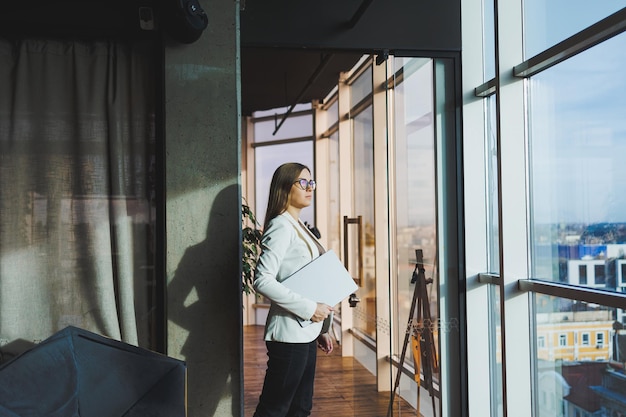 Una mujer de negocios con cabello largo en una chaqueta blanca y anteojos se para con una computadora portátil en sus manos en el espacio de la oficina cerca de la ventana y mira la ciudad Trabajar en una oficina moderna con grandes ventanales