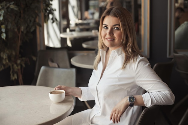 Mujer de negocios con camisa blanca de oficina con cabello rubio bebiendo una taza de café en un café