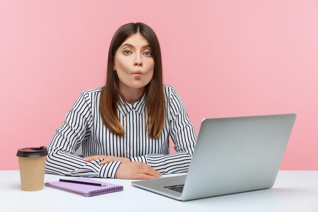 Mujer de negocios cansada divirtiéndose haciendo labios de pescado sentado en la computadora portátil en el lugar de trabajo haciendo una mueca ridícula sintiéndose culpable y confundida Disparo de estudio interior aislado sobre fondo rosa