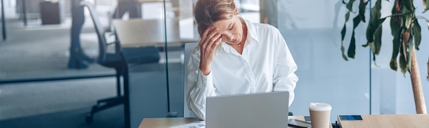 Mujer de negocios cansada con dolor de cabeza trabajando en una laptop en su lugar de trabajo en la oficina moderna