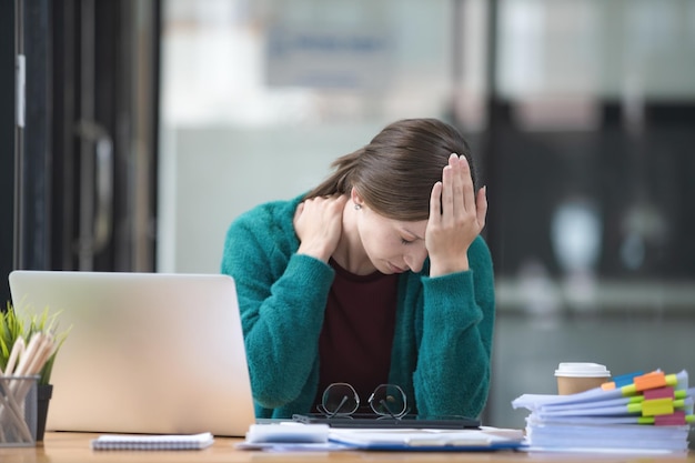 Una mujer de negocios cansada bajo estrés trabaja en una computadora portátil mientras está sentada en una mesa en casa y sostiene su mano en sus templos ataques de migraña