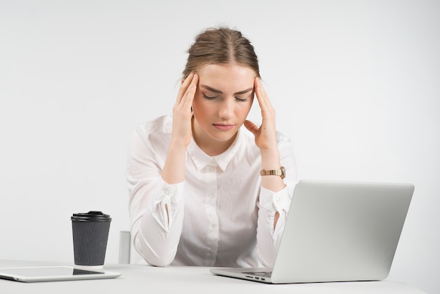 Mujer de negocios cansada que se sienta detrás de un ordenador portátil con una taza de café y de ipad en la tabla y que toca su cabeza para las manos