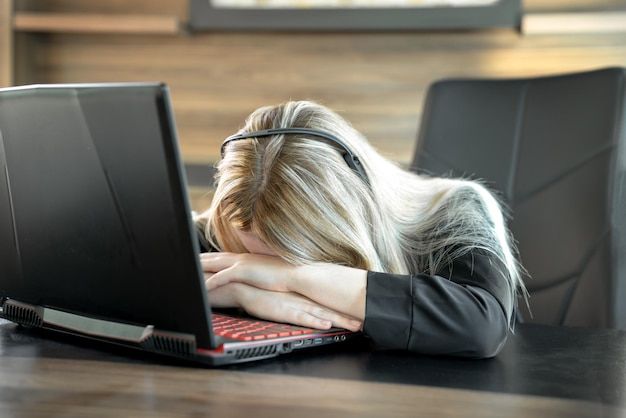 Mujer de negocios cansada en traje negro está sentada frente a la computadora portátil en la oficina cabeza está descansando en las manos