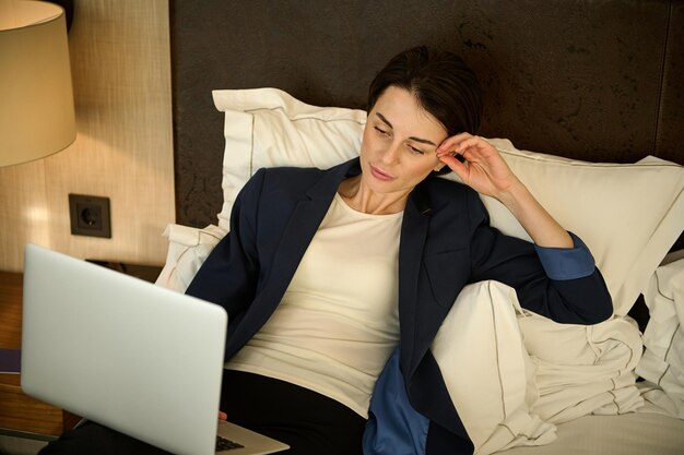 Mujer de negocios caucásica hermosa cansada pensativa en traje casual elegante que trabaja en la computadora portátil acostada en la cama, descansando después de un duro día de trabajo