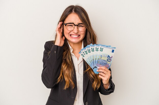 Mujer de negocios caucásica joven que sostiene los billetes aislados en el fondo blanco que cubre los oídos con las manos.