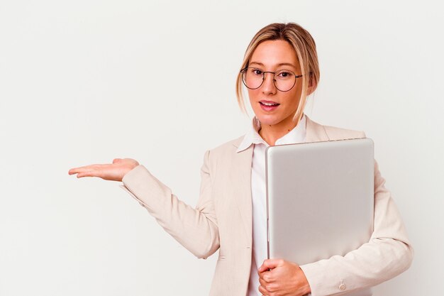 Mujer de negocios caucásica joven que sostiene una computadora portátil aislada en blanco que muestra un espacio de la copia en una palma y que sostiene otra mano en la cintura.