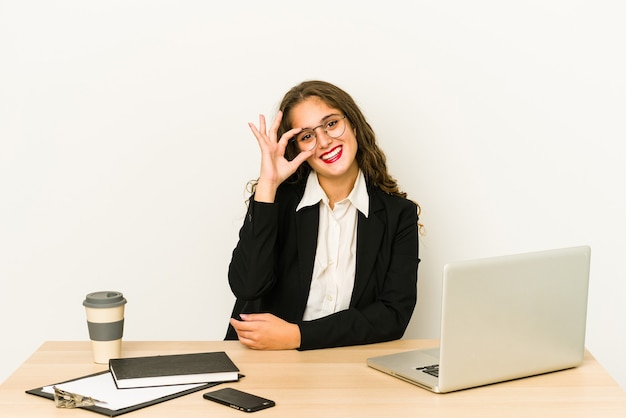 Mujer de negocios caucásica joven que trabaja en su escritorio aislado emocionado manteniendo el gesto ok en el ojo.