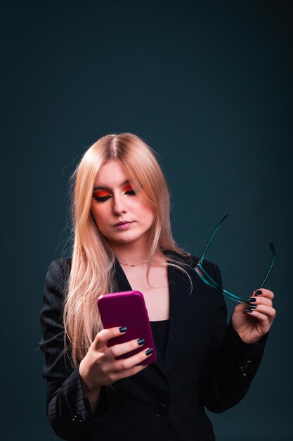 Mujer de negocios caucásica joven que usa un teléfono móvil que lleva un blazer negro y gafas en un estudio de fotografía con un fondo azul oscuro.