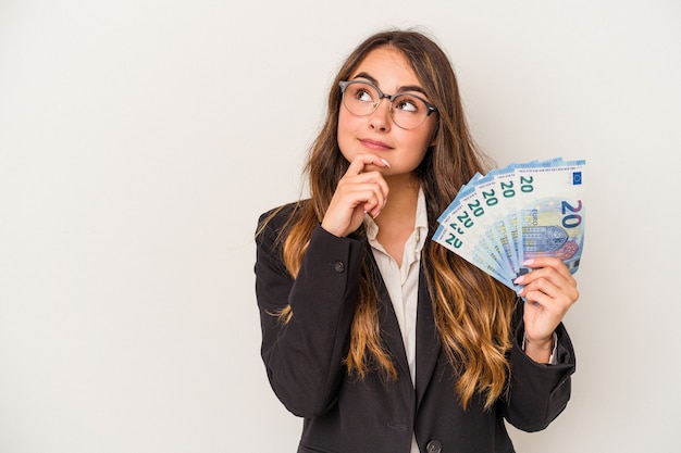 Mujer de negocios caucásica joven sosteniendo billetes aislados sobre fondo blanco mirando hacia los lados con expresión dudosa y escéptica.