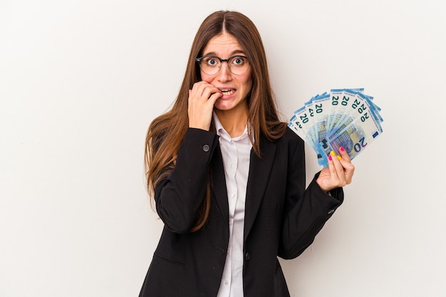 Mujer de negocios caucásica joven sosteniendo billetes aislados sobre fondo blanco mordiéndose las uñas, nerviosa y muy ansiosa.