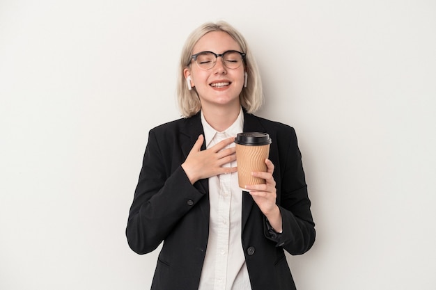 Mujer de negocios caucásica joven sosteniendo café para llevar aislado sobre fondo blanco se ríe a carcajadas manteniendo la mano en el pecho.