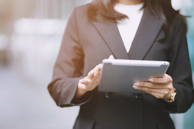 Mujer de negocios de cerca que trabaja con un dispositivo de tableta digital mientras está de pie frente a las ventanas en un edificio de oficinas con vistas a la ciudad.