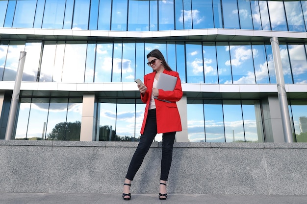 Una mujer de negocios en una chaqueta posa con una computadora portátil contra el fondo de los edificios