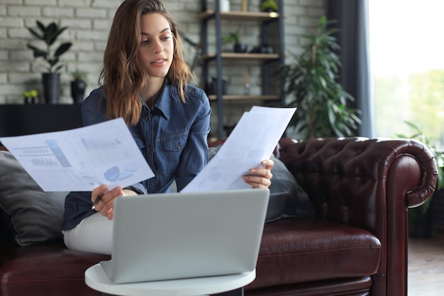 Mujer de negocios comprobando documentos en papel en la oficina en casa, trabajando en la computadora portátil desde casa.