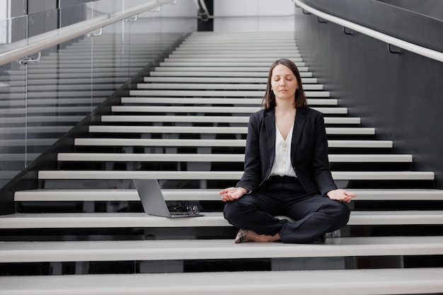 Mujer de negocios con una computadora portátil en la oficina del centro de negocios se sienta en los escalones en posición de loto y medita relajarse
