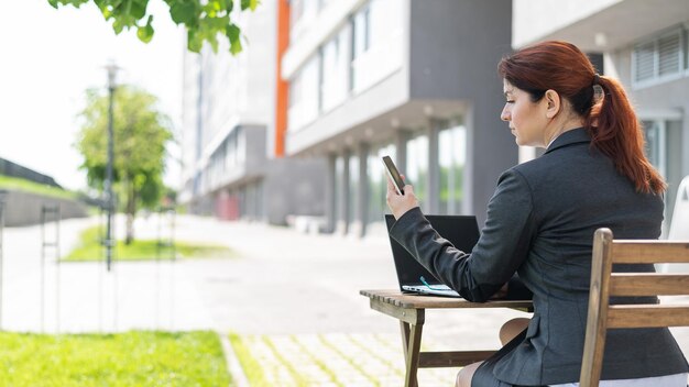 La mujer de negocios se comunica en su móvil sentada en una mesa con una computadora portátil en un café en la terraza de verano Vista trasera de una niña que trabaja al aire libre y hace una videollamada Escribiendo en el teléfono inteligente