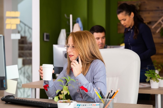 Mujer de negocios concentrada mirando en el monitor de su computadora mientras tiene una conversación telefónica. Mujer de negocios sosteniendo una taza de café.