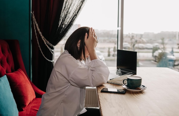 Una mujer de negocios deprimida está sentada en una mesa en un café con una computadora portátil y papeles